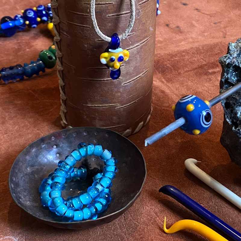 blaue Perlenkette in einem braunen Schälchen, daneben eine blaue Glasperle mit gelben Verzierungen, die noch auf einem Stab steck
