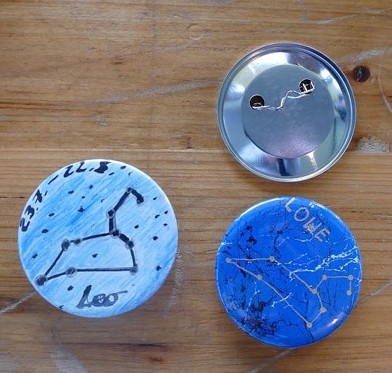 Drei Buttons liegen auf einem Holztisch, bei einem ist die Rückseite sichtbar. Bei zweien ist die Vorderseite zu sehen, auf denen Sternbilder aufgemalt wurden. 