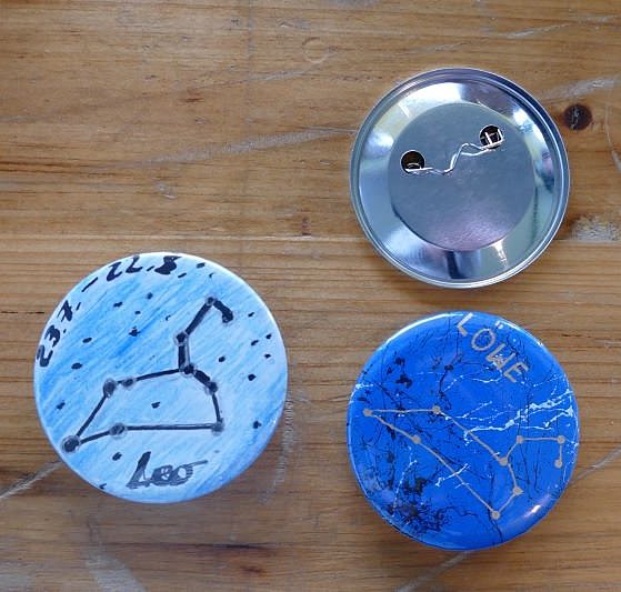 Drei Buttons liegen auf einem Holztisch, bei einem ist die Rückseite sichtbar. Bei zweien ist die Vorderseite zu sehen, auf denen Sternbilder aufgemalt wurden. 