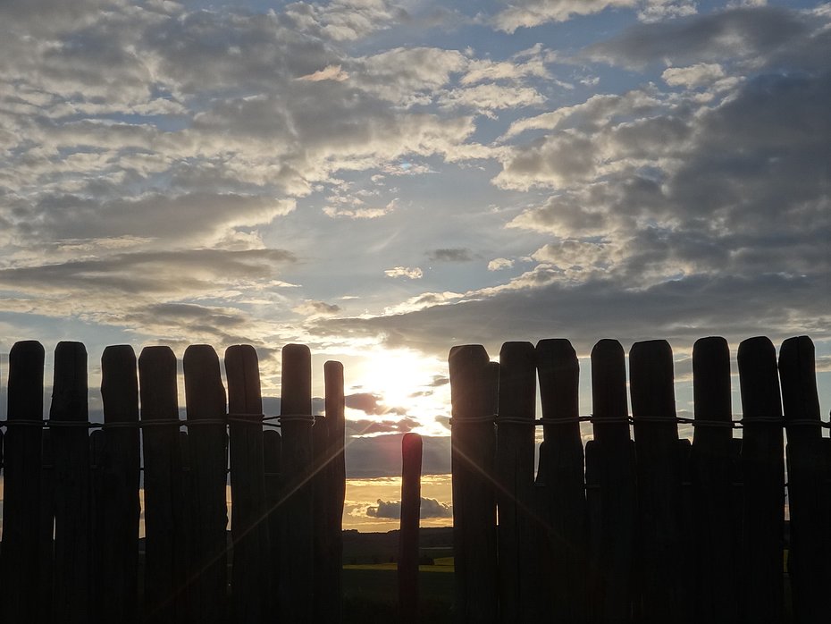 Ein Abendhimmel mit Wolken, im Vordergrund schattenhaft eine Reihe aus unregelmäßigen Pfosten. In der Bildmitte ist eine Lücke zwischen den Pfosten, durch die die untergehende Sonne zu erkennen ist. 